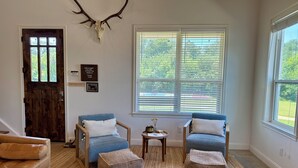 A cozy living room with a wooden door, two blue armchairs, a brown leather couch, and a large window with blinds. - Close to Baylor & Silos: 3 BR Home Pickleball Court, Ping Pong Table & Barn (Lorena)