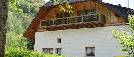 Eine zweistöckige Holzhütte mit einem Balkon, umgeben von Grünpflanzen und einer Steinmauer.