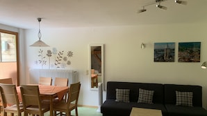 A living room with a black sofa, wooden chairs, a small table, and a dining table with chairs. There are wall decorations and a radiator.