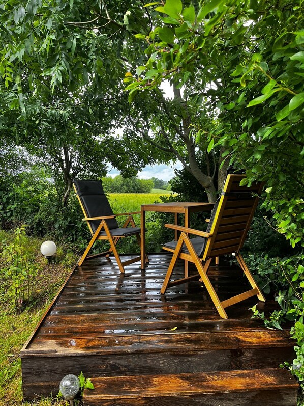 A wooden deck with two folding chairs and a table, surrounded by lush greenery and a view of a field.
