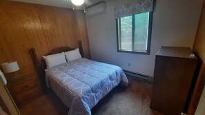 Une chambre à coucher avec une tête de lit en bois, un lit recouvert d’une literie à motifs, une table de chevet avec une lampe, une fenêtre avec des stores et une commode en bois.