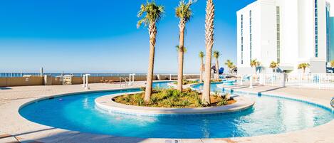 A swimming pool with palm trees, a tiled deck, and a hotel building in the background.