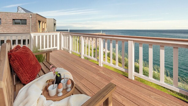 A wooden deck with a white railing overlooking a body of water, a breakfast setup with eggs and a drink, and a red cushion. - Land`s End - Blick auf die Freie Ostsee, Sauna, Wintergarten, Elektro Grill (Olpenitz)