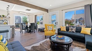 A spacious living room with a large sofa, a yellow armchair, and a glass coffee table. There is a dining area with a table and chairs, and a kitchen area with a counter and appliances.