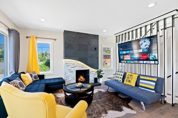 A modern living room with a stone fireplace, a large flat-screen TV, a blue sofa, and a yellow armchair. - 5-bedroom vacation home just 7 minutes to T-Mobile Park (Seattle)