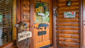 A wooden cabin entrance with a bear sculpture holding a 'Welcome' sign, a 'No Smoking' sign, a 'Bare Paws Please' sign, a 'Welcome' sign above the door, and a 'We're Bearly Hangin' On' sign below the door. - "Shine Valley #53" View for days (sevierville)
