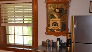 A kitchen with wooden cabinets, a stainless steel refrigerator, a window with blinds, and a framed picture above the sink. - Enderly - c1859 - English Basement Apartment (Charlottesville)