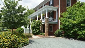 A two-story brick building with a white porch and columns, surrounded by greenery and a gravel driveway. - Enderly - c1859 - English Basement Apartment (Charlottesville)