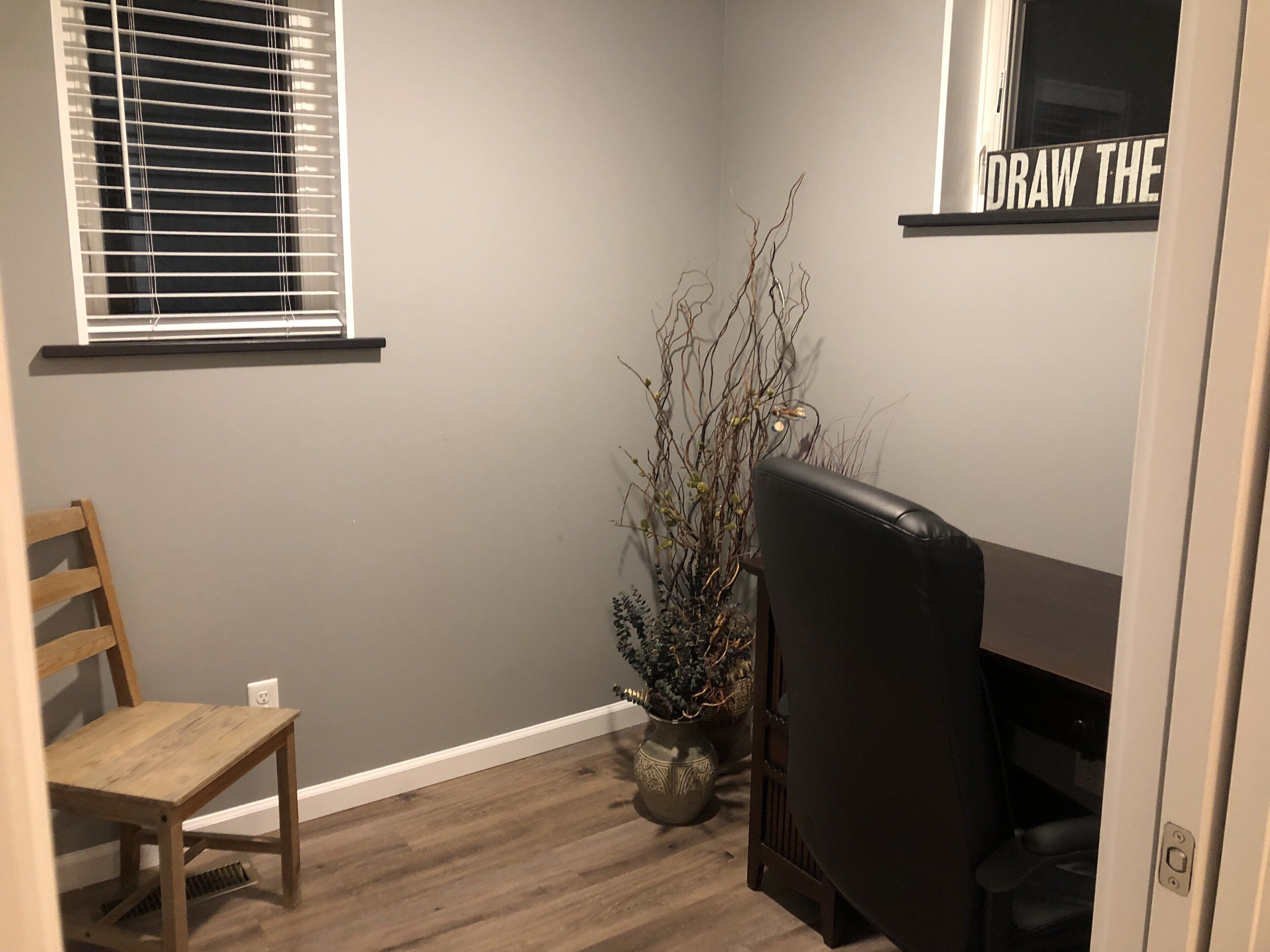 Una habitación de esquina con una silla de madera, una silla de oficina negra, una planta en maceta y una ventana con persianas.