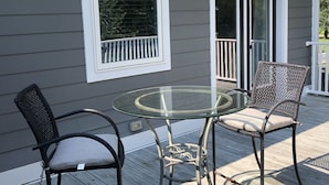 A glass-top table with two wicker chairs on a wooden deck, adjacent to a house with a white window and a door. - Newly Constructed Second Floor Modern Studio Apartment Unit 2 ~ Sleeps 6 (Accomac)