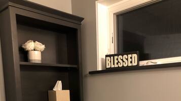 Un baño con inodoro, un estante con una caja de pañuelos y un florero, y una ventana con un letrero que dice "BLESSED".