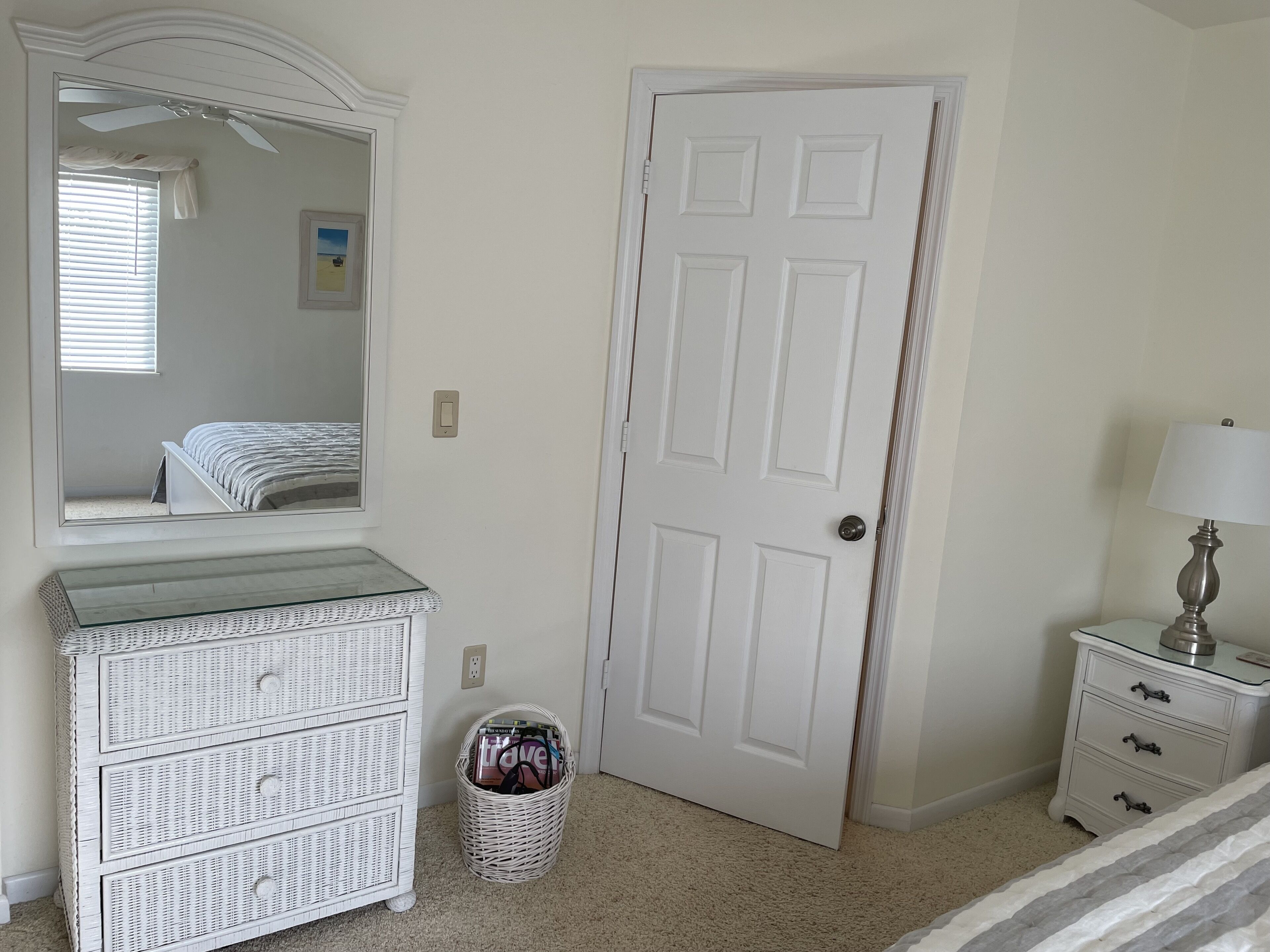 A bedroom with a white dresser, a mirror, a door, a lamp, and a basket.