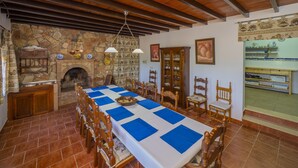 Une salle à manger avec une longue table, des chaises, une cheminée en pierre et un coin cuisine visible à travers une porte ouverte.