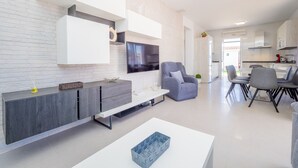 Ein modernes Wohnzimmer mit weißer Backsteinwand, einer grauen Couch, einem Flachbildfernseher und einem Essbereich mit grauen Stühlen und einem weißen Tisch.