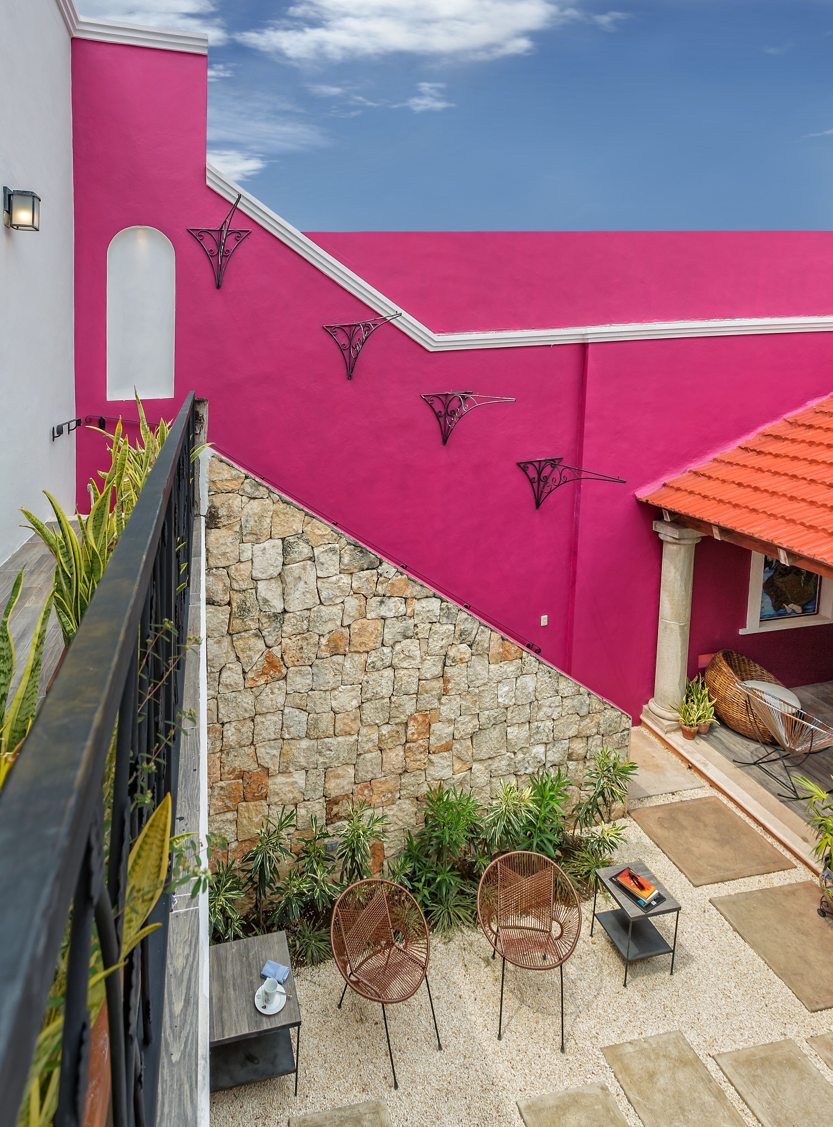 Una pared exterior rosa vibrante con detalles metálicos decorativos, una pared de piedra y un área de descanso con sillas de mimbre y una mesa.