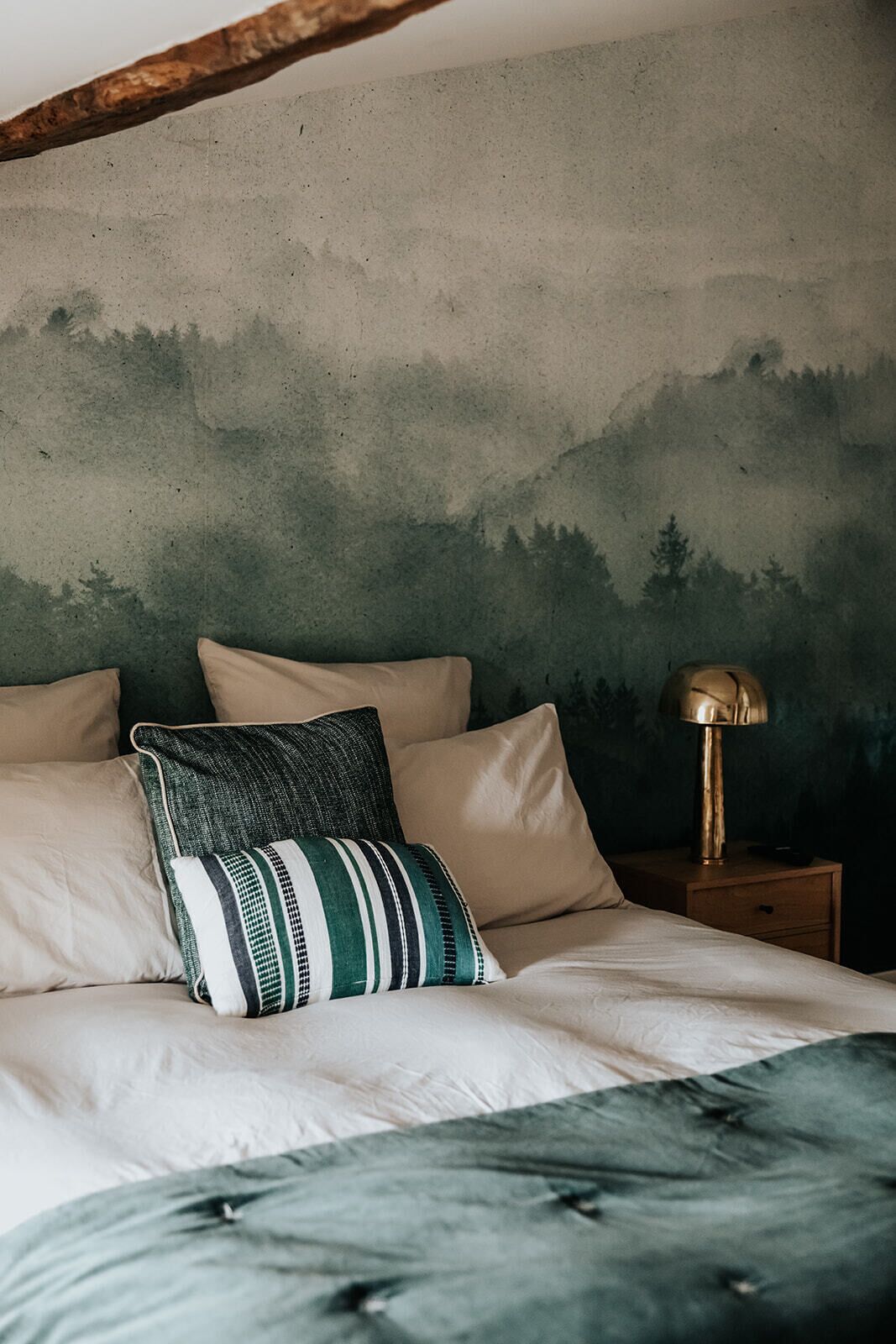 Een bed met een kussen met groene en witte strepen, een beige kussen en een groen kussen. Er staat een nachtkastje met een lamp erop.