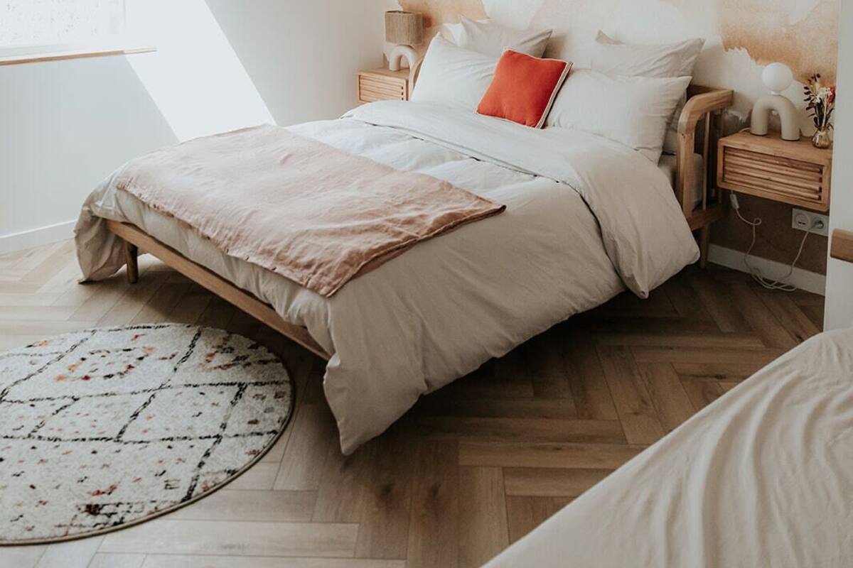 A bedroom with a bed, a round rug, a wooden headboard, a nightstand, and a wall with a mountain painting.