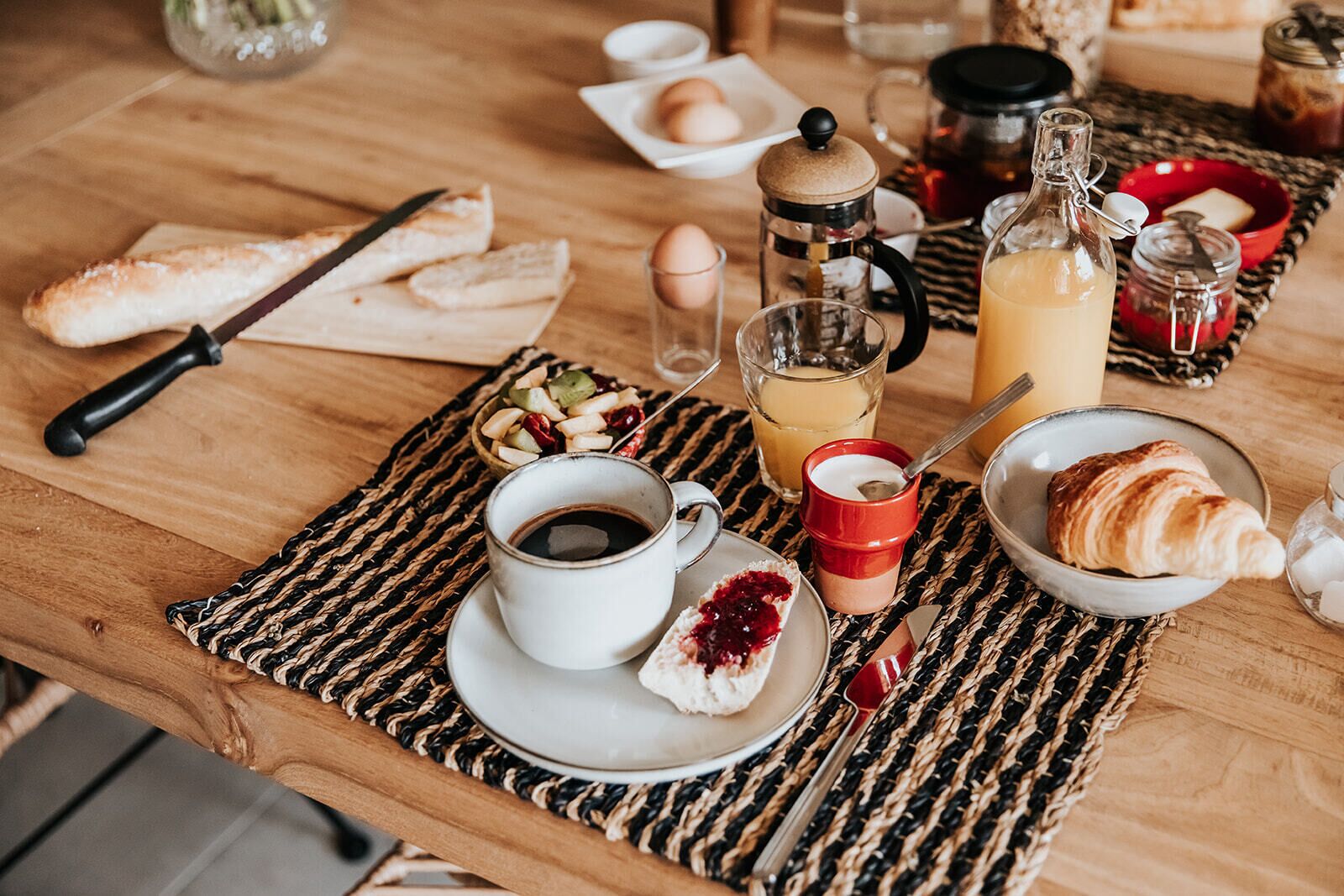 Un desayuno con pan, croissants, una taza de café, un vaso de zumo de naranja y un bol pequeño de fruta.