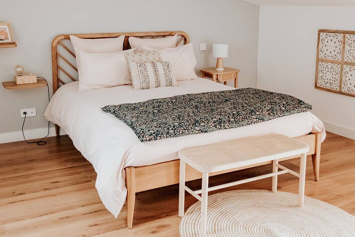 A bedroom with a wooden bed, a woven bench, a round rug, and a wooden ceiling beam.