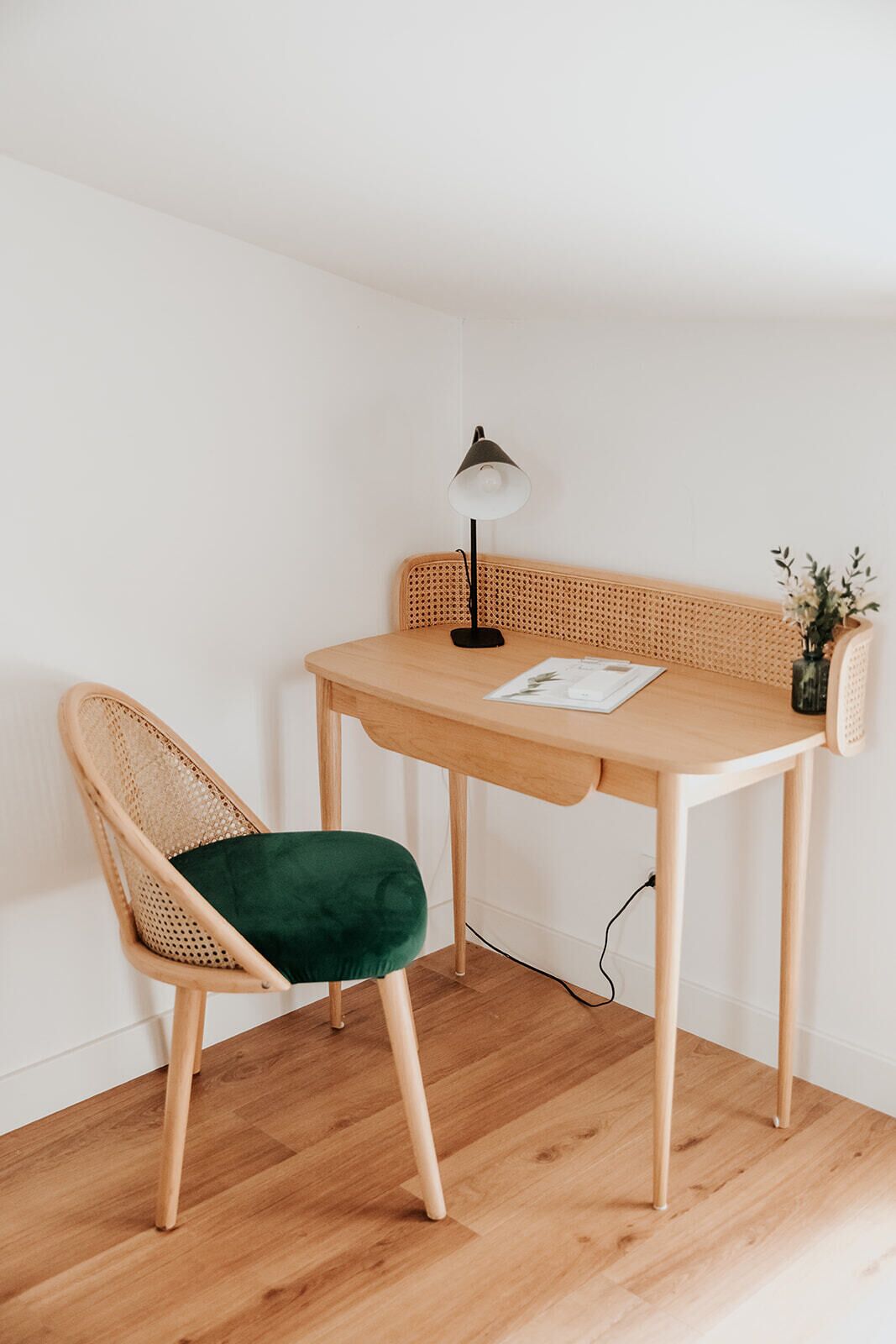 Una habitación de esquina con un escritorio de madera, una silla de mimbre con cojín verde, una lámpara de escritorio negra y una planta pequeña sobre el escritorio.