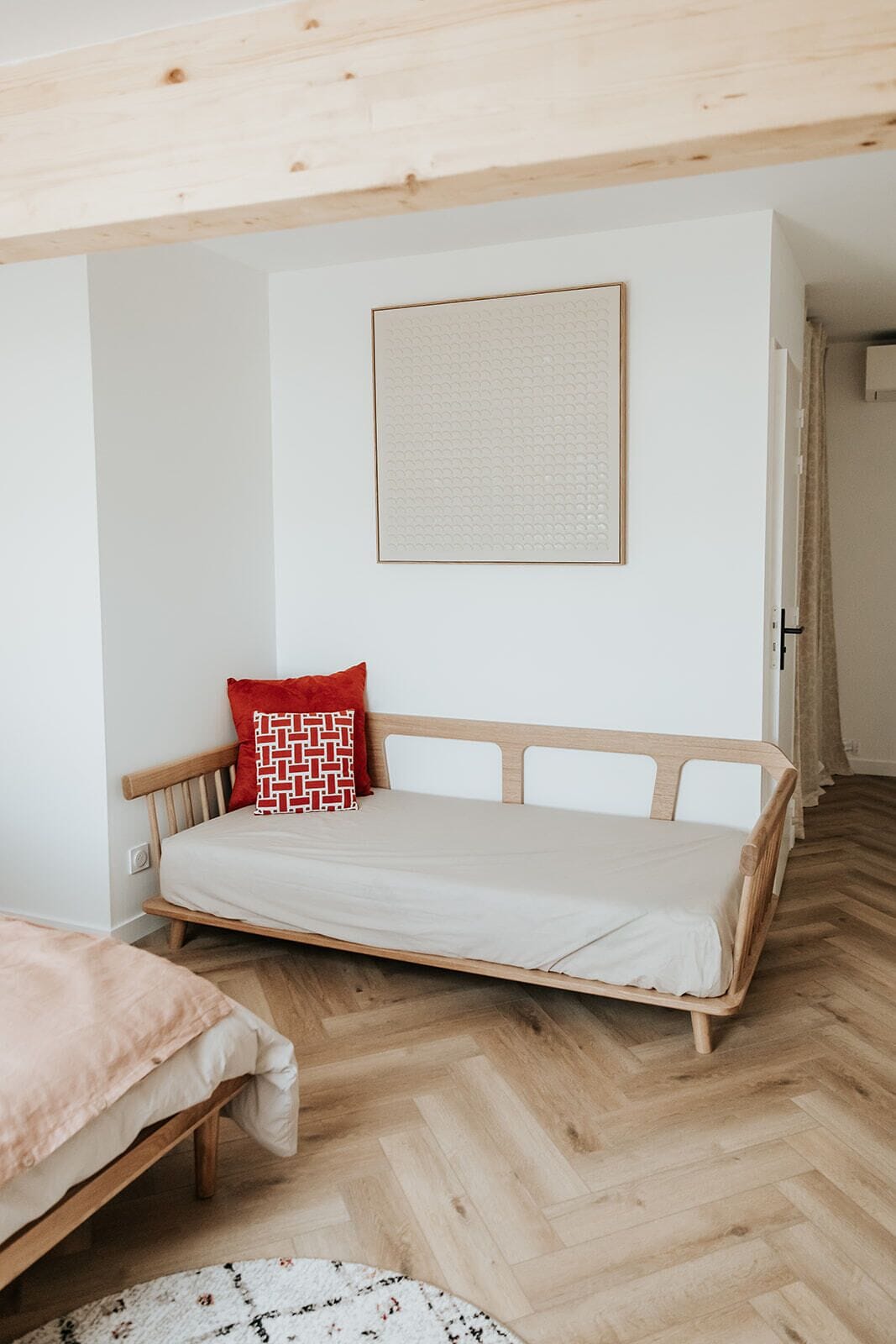 Una habitación con una cama de madera, un almohadón rojo, una pared blanca y un cuadro enmarcado.