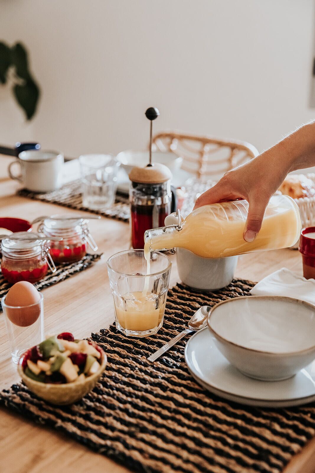 Un desayuno servido con una mano vertiendo jugo de naranja en un vaso, un bol de fruta y diferentes alimentos para el desayuno sobre un mantel tejido.