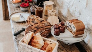 A rustic wooden table with various food items, including bread, pastries, and condiments, set against a white stone wall. - Château La Grande Clotte (Lussac)