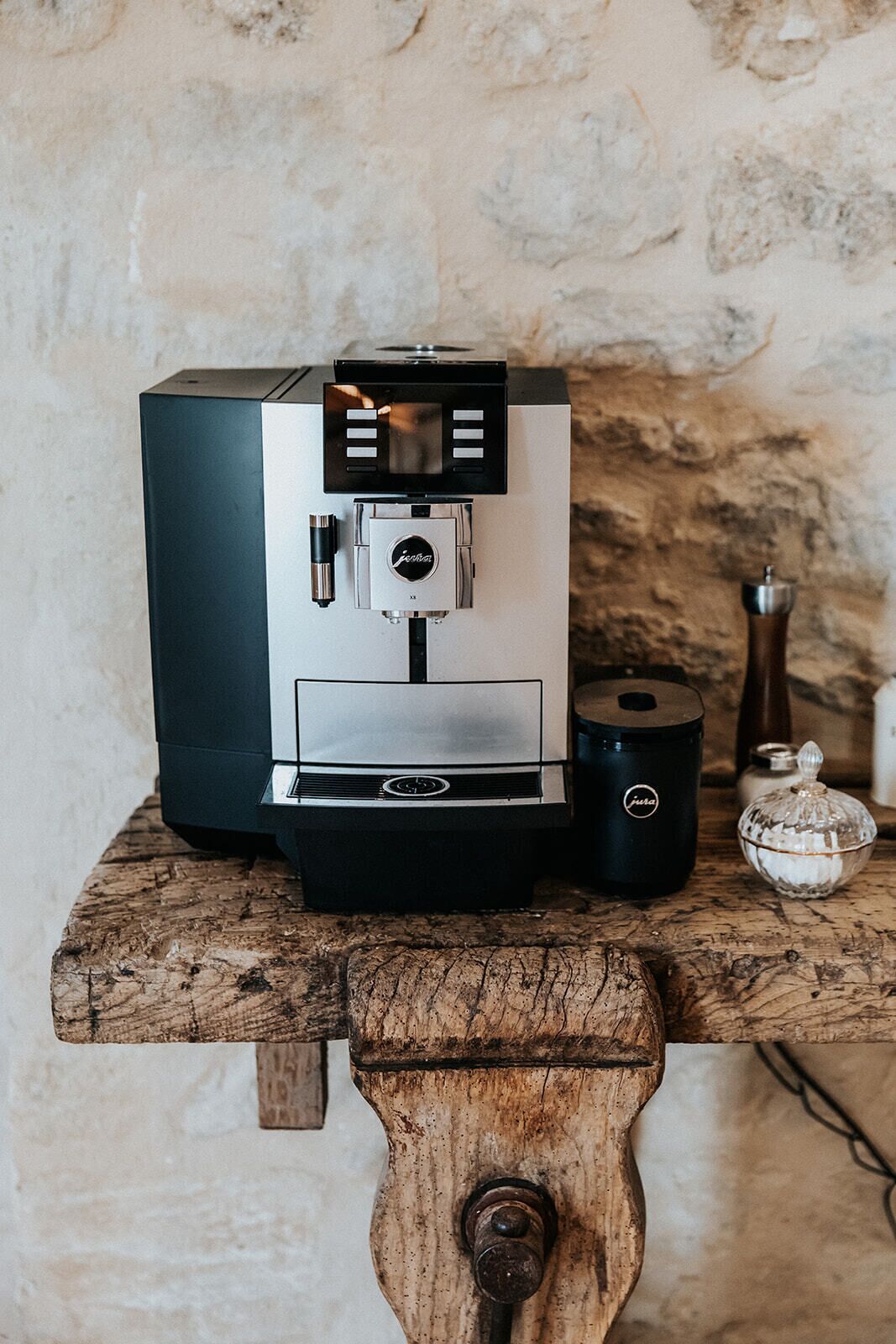 Máquina de café sobre uma mesa de madeira rústica, com parede de pedra ao fundo.