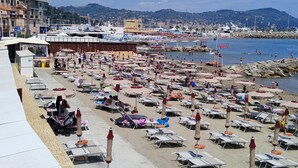 A beach with numerous sun loungers and umbrellas, a marina in the background, and buildings along the coastline.