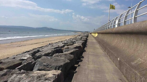 Ein Küstenweg mit einer Betonbarriere, einem Sandstrand und dem Meer im Hintergrund.