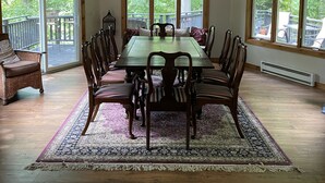 A dining room with a large wooden table, six chairs, a chandelier, and large windows with blinds.