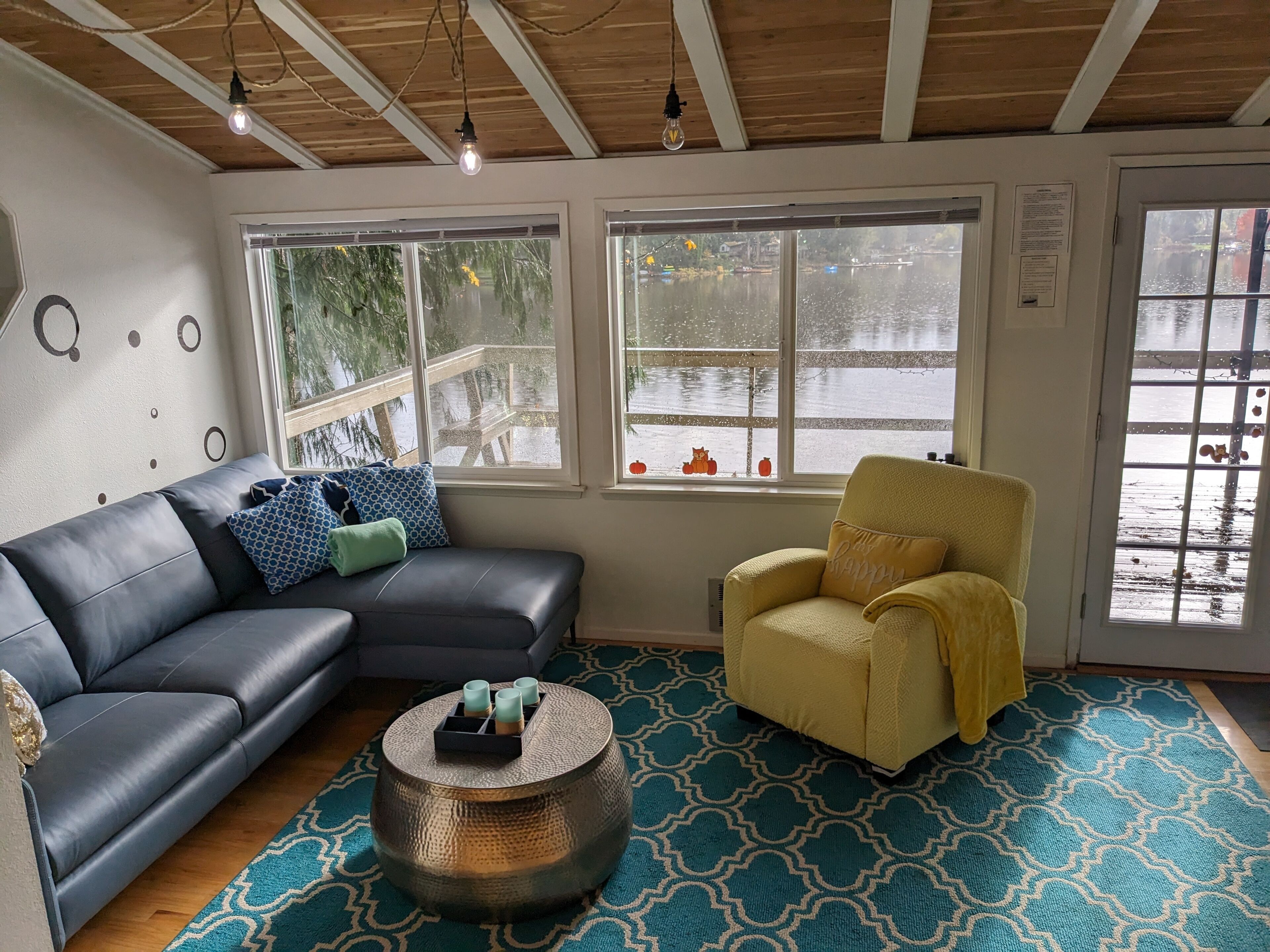 A cozy living room with a black leather sofa, a yellow armchair, and a round coffee table with a tray of candles. Large windows offer a view of a wooden deck and a body of water.