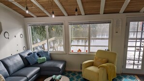 Un salon chaleureux avec un canapé en cuir noir, un fauteuil jaune et une table basse ronde sur laquelle est posé un plateau avec des bougies. De grandes fenêtres offrent une vue sur une terrasse en bois et un plan d’eau.