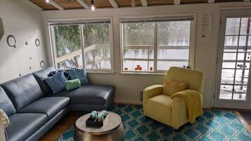 A cozy living room with a black leather sofa, a yellow armchair, and a round coffee table with a tray of candles. Large windows offer a view of a wooden deck and a body of water.
