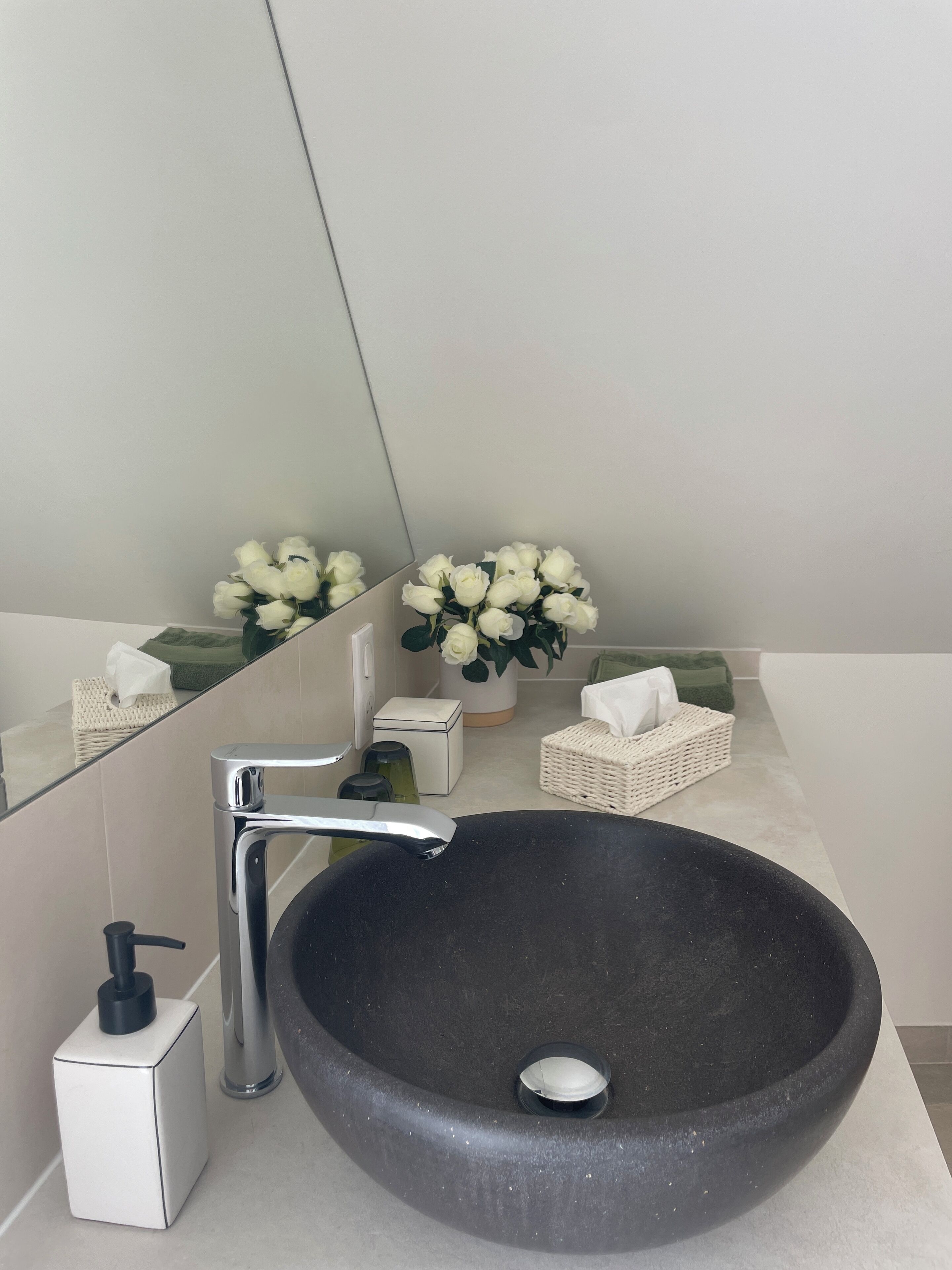 A modern bathroom with a large vessel sink, a mirror, a white soap dispenser, a small white box, a potted white rose, and a wicker basket.