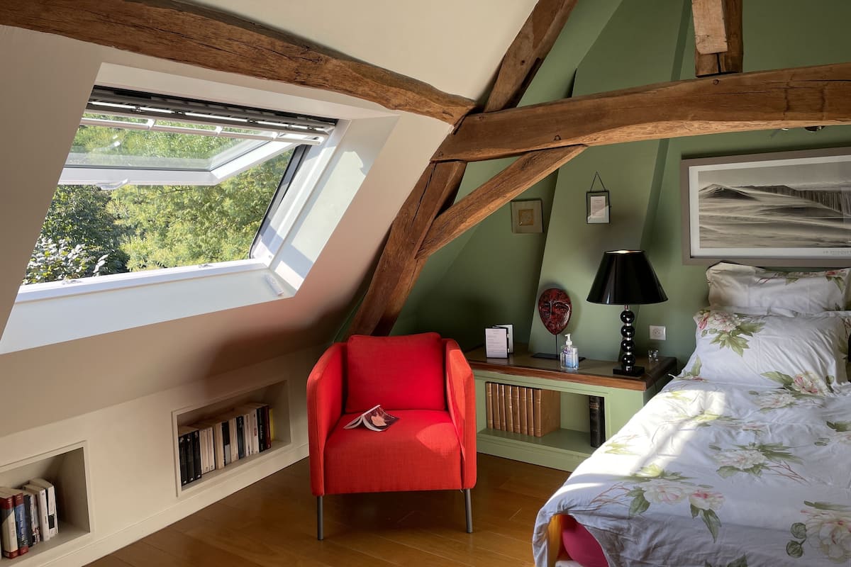 A room with a sloped ceiling, a red chair, a bed with floral bedding, a desk with a lamp, and a framed picture on the wall.