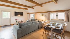A cozy living room with a grey sofa, wooden beams, a dining table set for four, and a TV mounted on the wall. - Motor House Loft - beautiful loft conversion in lovely rural location by Bloom Stays (Sandhurst)