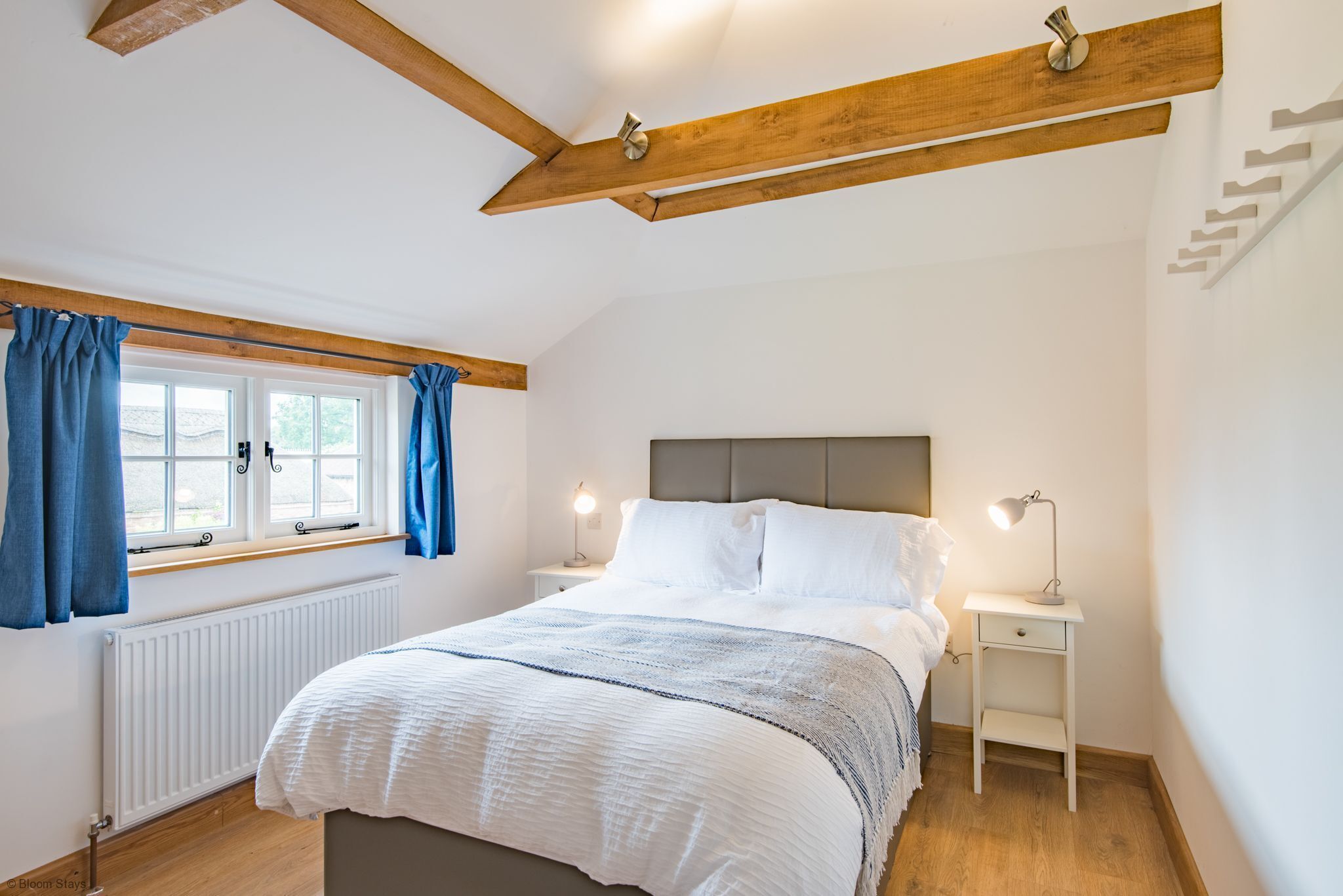 Quarto com cama, cortinas brancas, janela, mesa de cabeceira com luminária e radiador.