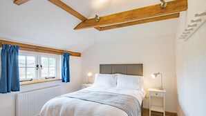 Quarto com cama, cortinas brancas, janela, mesa de cabeceira com luminária e radiador.