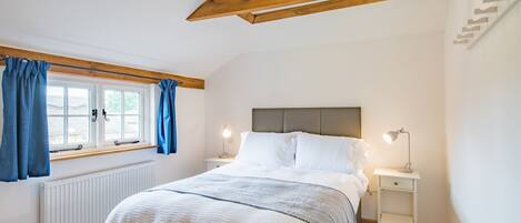 Quarto com cama, cortinas brancas, janela, mesa de cabeceira com luminária e radiador.