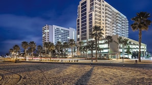 Un hotel frente a la playa con varios pisos, palmeras y una playa de arena.