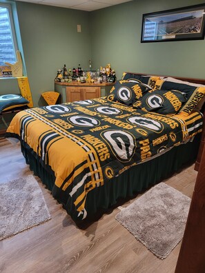 Quarto com cama com colcha temática de equipe esportiva, cômoda com diversos objetos e janela com vista para um estádio.