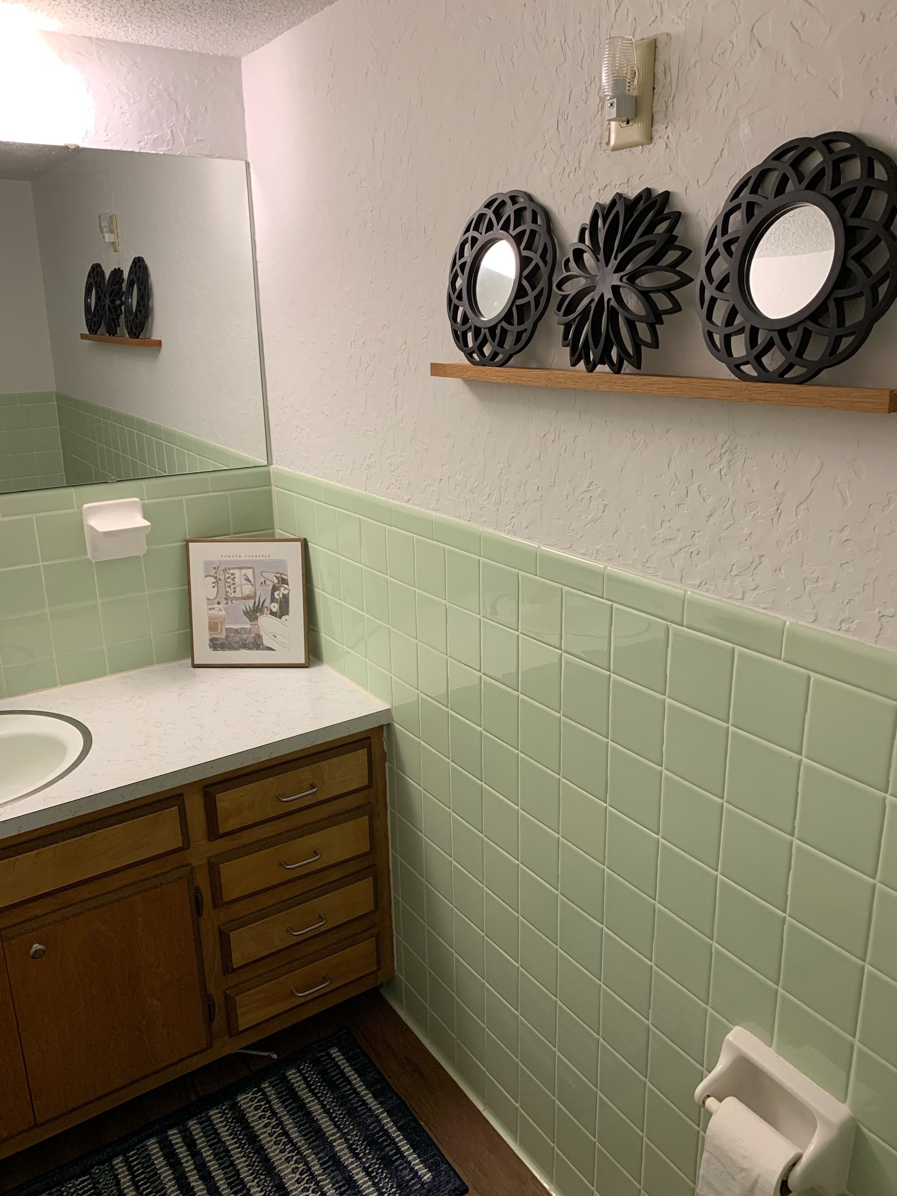 A bathroom with a white sink, wooden vanity, green-tiled walls, a mirror, decorative wall pieces, and a framed picture on the countertop.