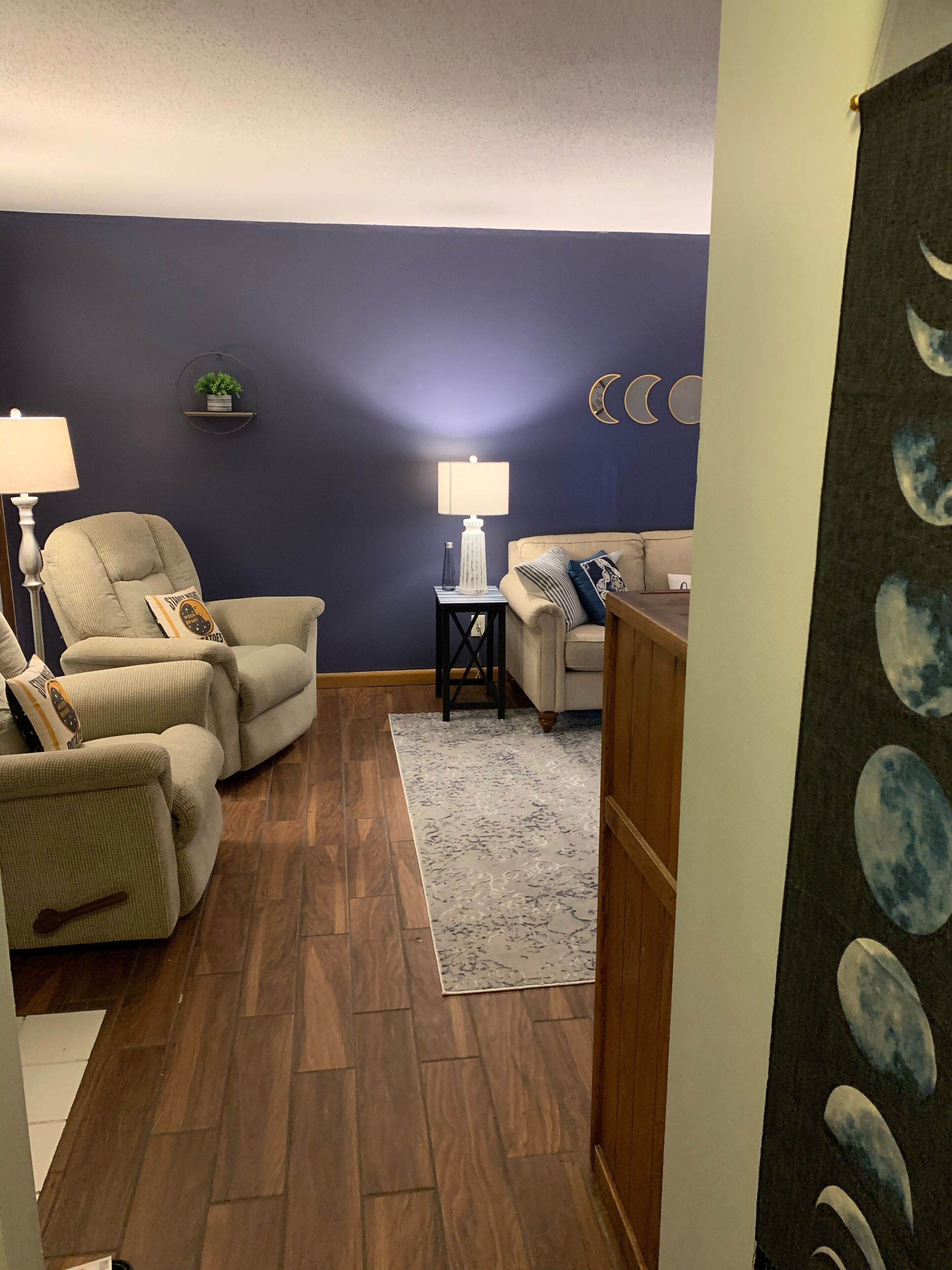 Una sala de estar con una pared azul oscuro, un sofá color beige, un sillón blanco, una mesita auxiliar con una lámpara y una alfombra.
