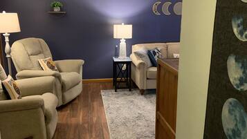 Una sala de estar con una pared azul oscuro, un sofá color beige, un sillón blanco, una mesita auxiliar con una lámpara y una alfombra.