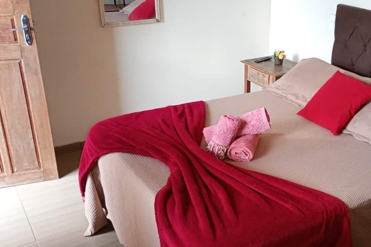 Un lit bien fait, recouvert d’une couvre-lit rouge et beige, avec un oreiller rouge et une table de chevet en bois ornée d’un vase de fleurs.