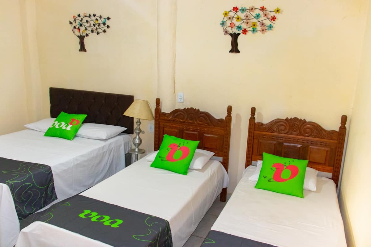 Une chambre d’hôtel avec trois lits simples, chacun doté d’une tête de lit en bois et d’un oreiller vert orné d’un logo « D » rouge.
