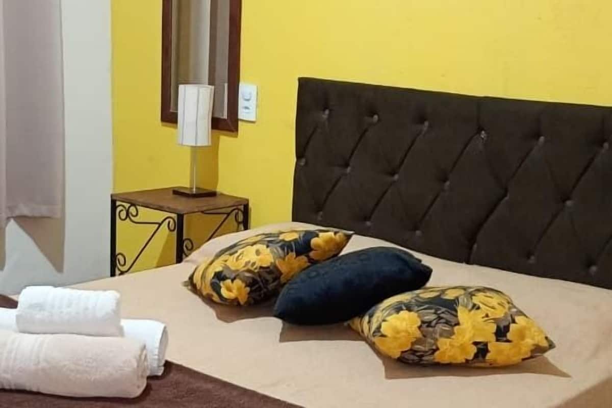 Une chambre à coucher avec un lit, des oreillers, une couverture, une table de chevet avec une lampe, un miroir et une œuvre d’art murale.