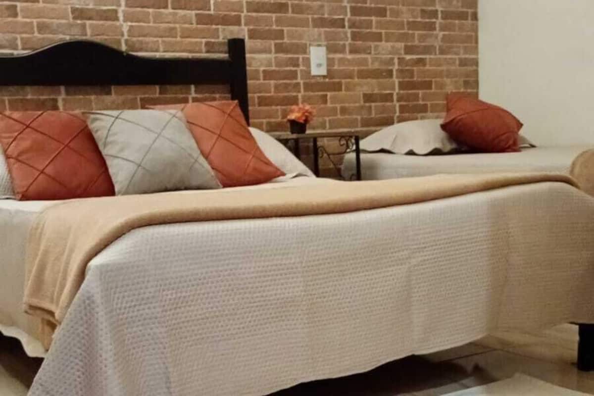 Un lit recouvert d’une couvre-lit blanche, avec une tête de lit en bois et un mur en briques derrière.