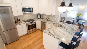 Una cocina moderna con armarios blancos, electrodomésticos de acero inoxidable, encimera de mármol y un comedor con una mesa redonda y sillas.